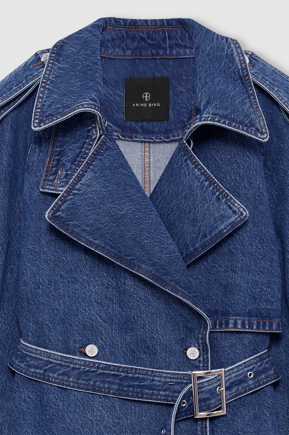 Close-up of the Pascale Trench by Anine Bing, a blue denim, military-inspired trench coat featuring wide lapels, silver buttons, a buckle belt, and a black ANINE BING label on the collar.
