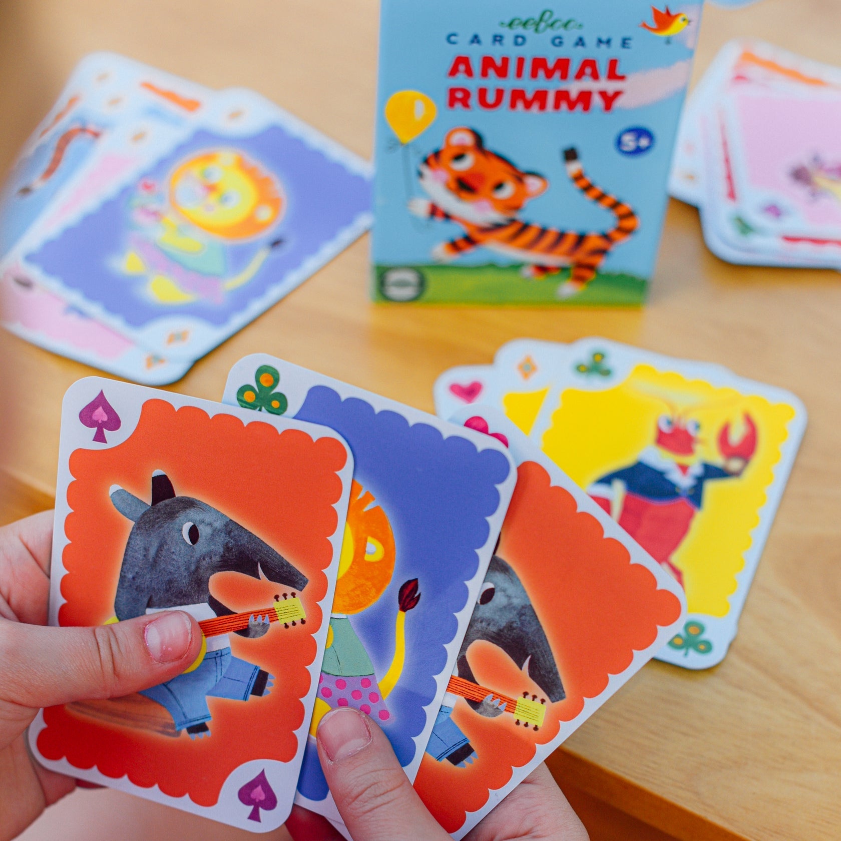 A person holds playing cards from the Eeboo Animal Rummy Playing Cards set, highlighting its vintage design with colorful animal illustrations. The game box and additional cards are on the wooden table, contributing to the nostalgic charm of this playful scene.