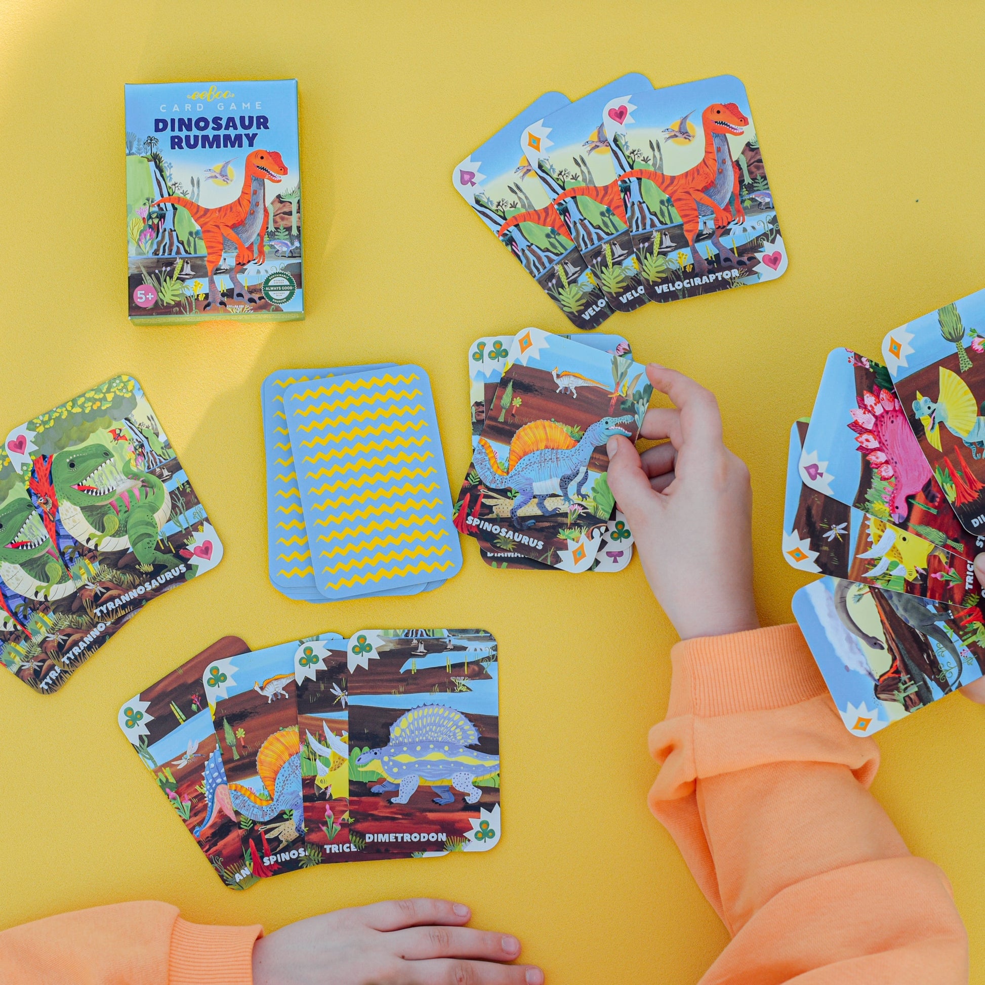 Wearing an orange sweatshirt, a person enjoys a fun and family-friendly game of Eeboo’s Dinosaur Rummy Playing Cards with an illustrated deck on a yellow surface.