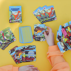 Two hands arrange Eeboo Dinosaur Rummy Playing Cards on a yellow surface, with the family-friendly game box and extra cards from the deck spread out nearby.