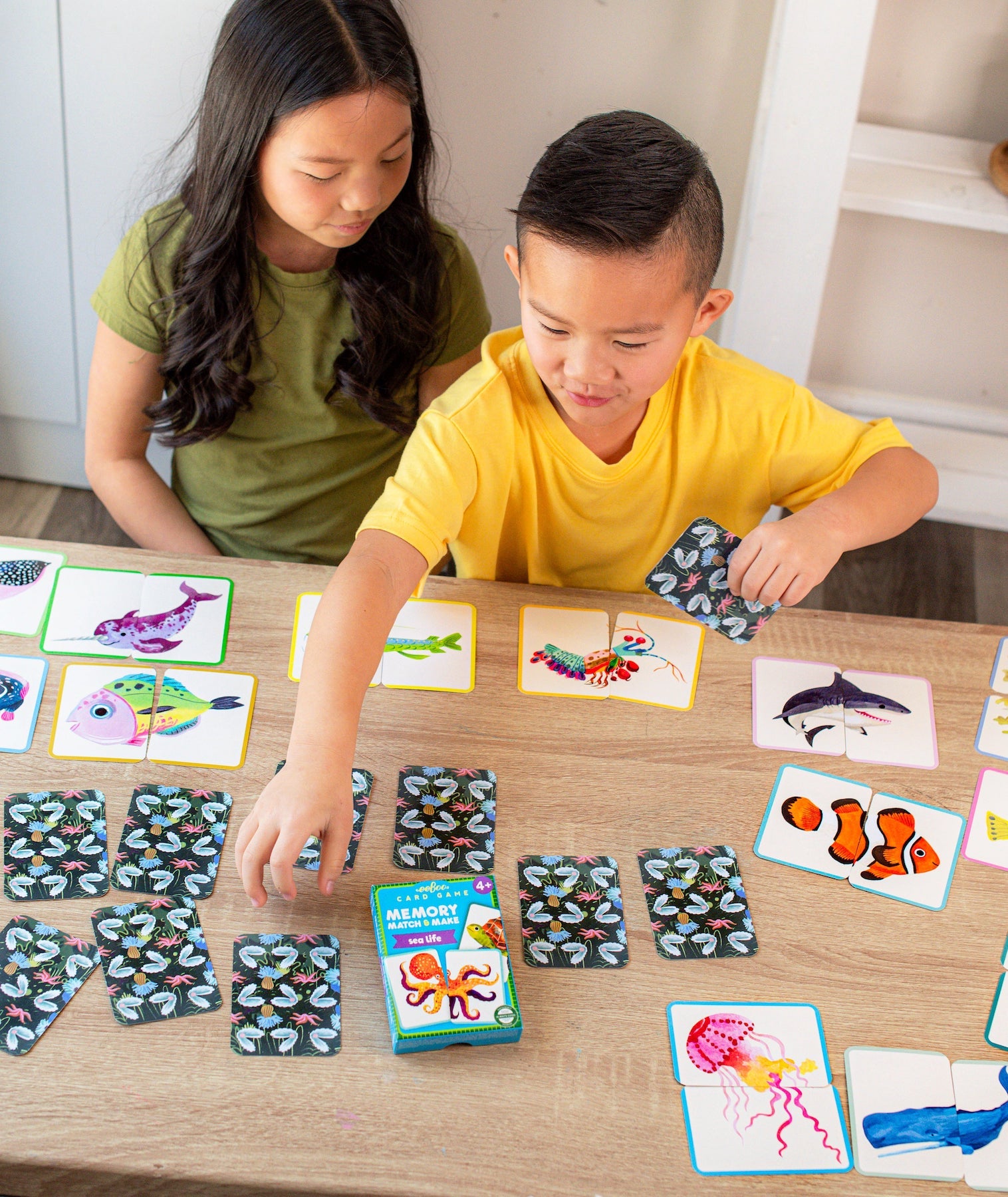 Two children play the Eeboo Memory Match & Make Card Game at a wooden table, enjoying matching cards that feature colorful illustrations of sea animals.