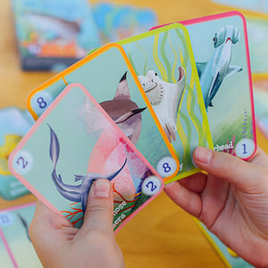 A person holds four illustrated cards featuring sea animals like a narwhal, shark, stingray, and hammerhead shark from Eeboo's family-friendly Go Shark Go! Playing Cards game.