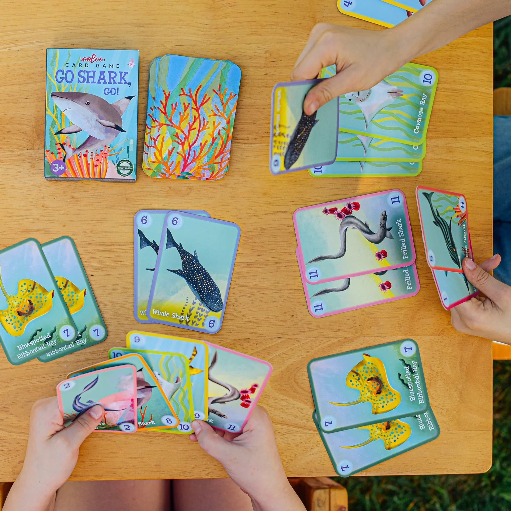 Four people are playing Go Shark Go! Playing Cards by Eeboo, a family-friendly shark-themed card game and Go Fish adaptation, on a wooden table with colorful sea creature cards spread out.