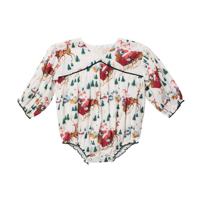 Pink Chicken Baby Girls’ Wynne Bubble long-sleeved holiday romper featuring a Santa sleigh print with reindeer, pine trees, and small houses, finished with dark green trim.