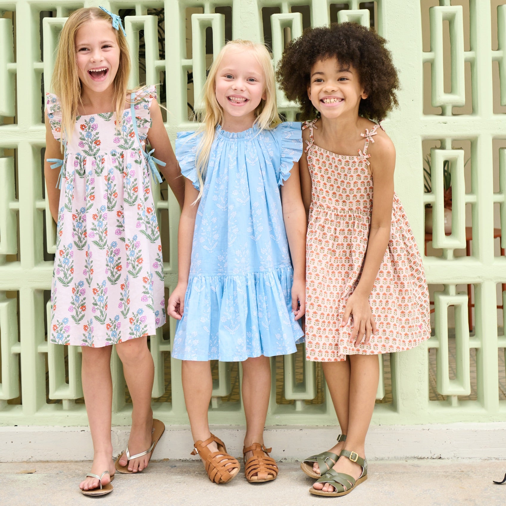 Three young girls stand side by side, smiling in Pink Chicken Girls' Harper Dresses and sandals, in front of a light green patterned wall.