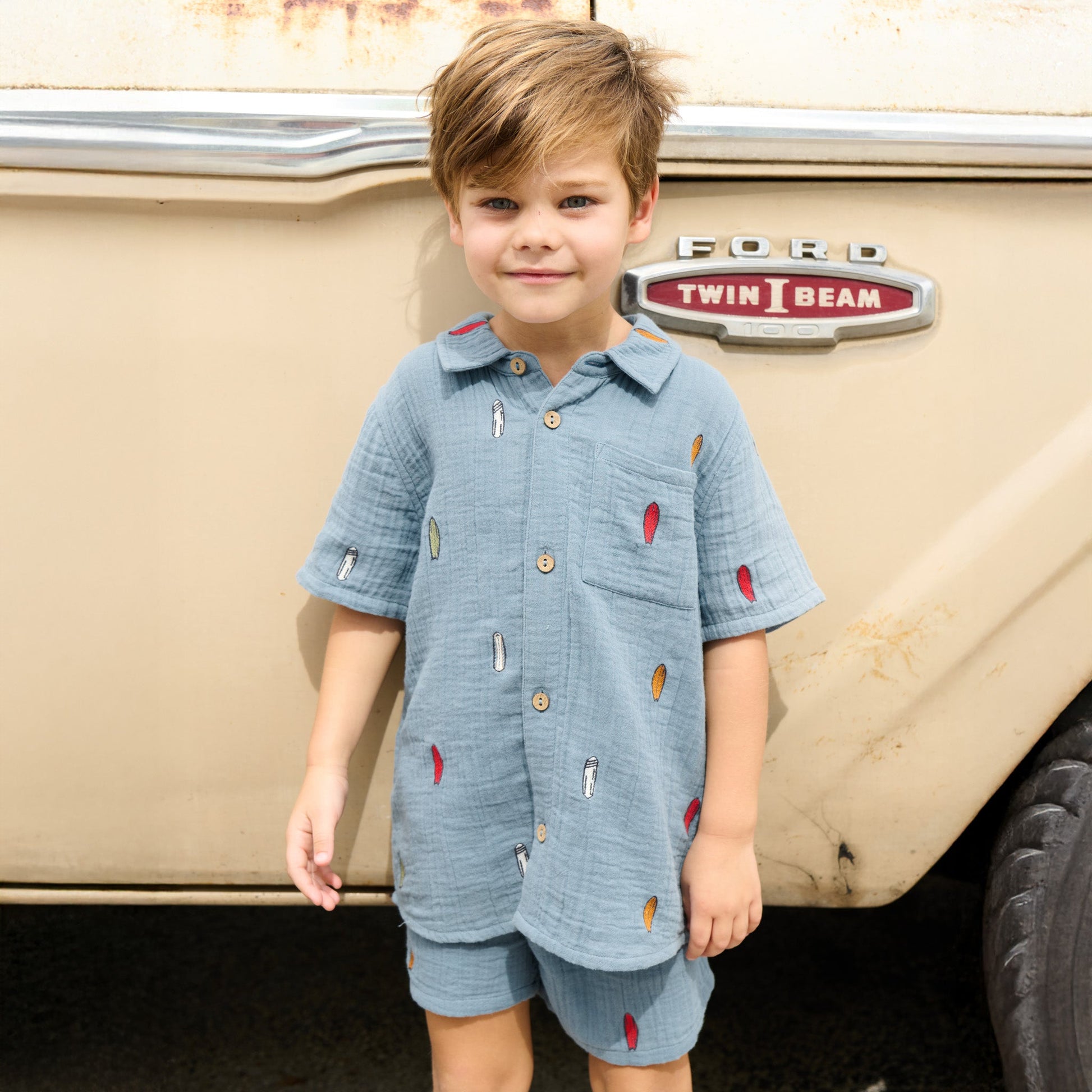 A young child wearing a blue shirt and Pink Chicken Boys’ Calvin Short stands in front of a beige Ford Twin I Beam vehicle.