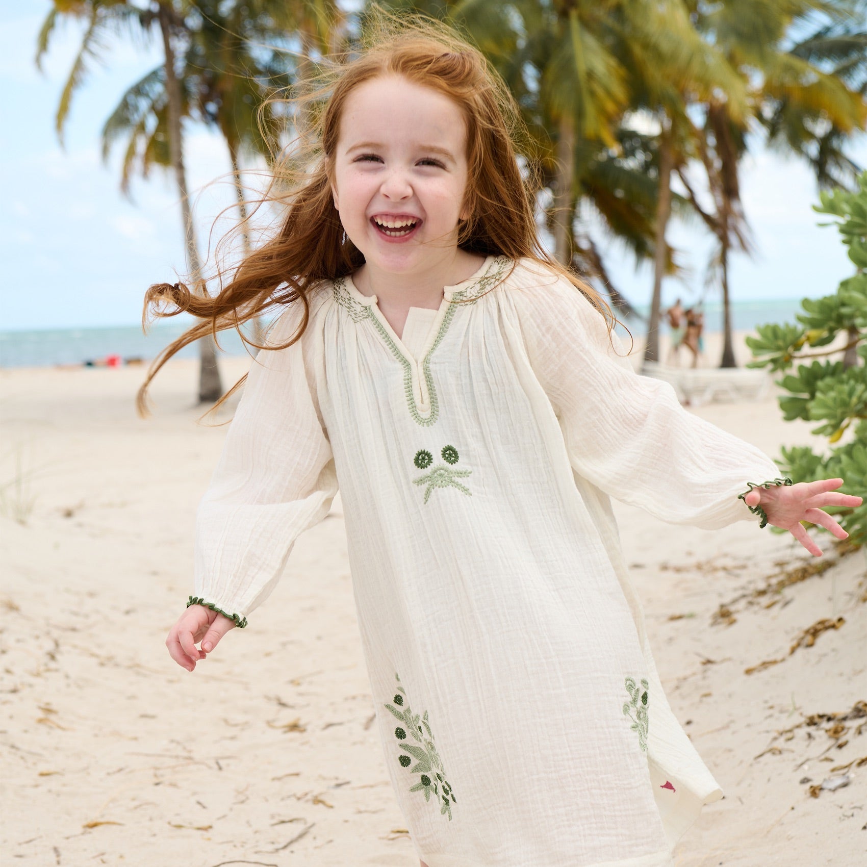 A young girl smiles while running on a sandy beach in the Girls' Ava Dress by Pink Chicken, featuring green embroidery and soft cotton gauze fabric that flutters as she moves among the palm trees.