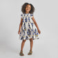 A young girl smiles in the Pink Chicken Girls' Stevie Dress—Navy Buttercup Floral with hand-smocked details, paired with green sandals, standing against a plain light background.