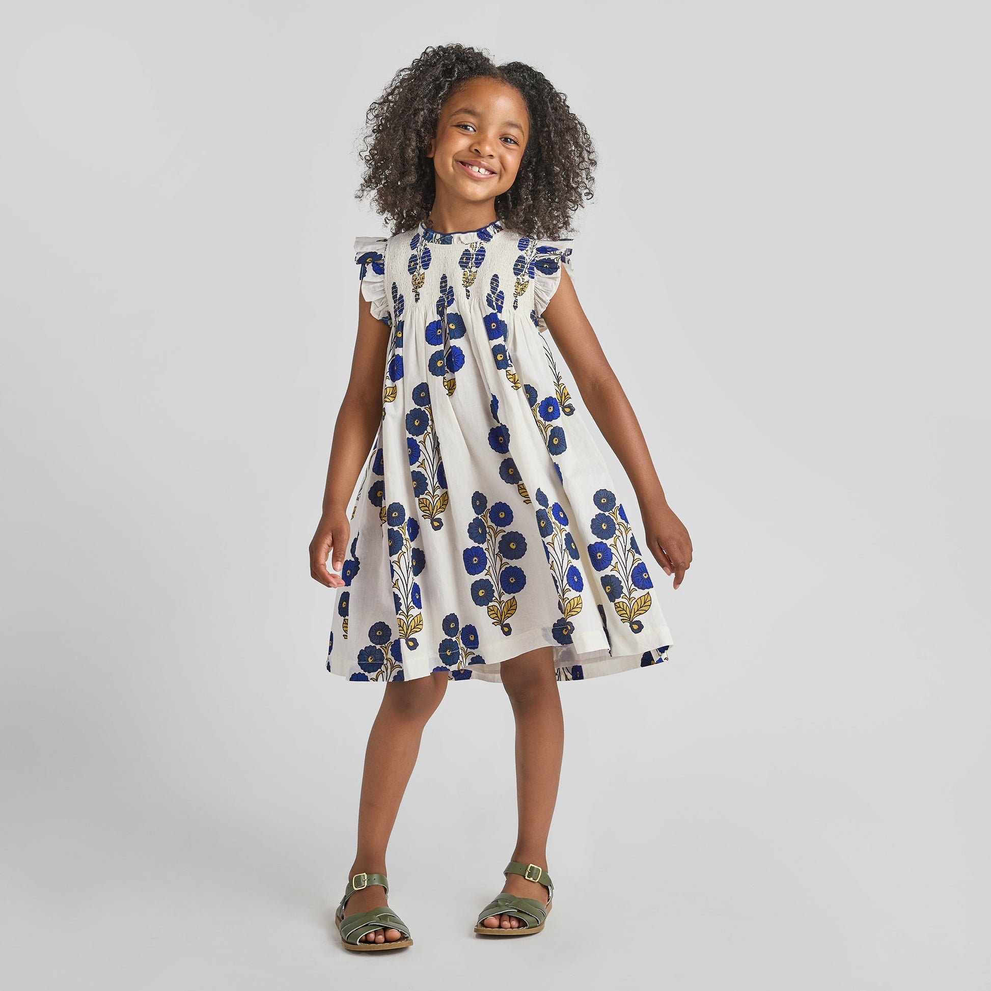 A young girl smiles in the Pink Chicken Girls' Stevie Dress—Navy Buttercup Floral with hand-smocked details, paired with green sandals, standing against a plain light background.