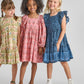 Three girls stand side by side in Pink Chicken Girls' Elsie Dresses, each featuring a charming block print—yellow, pink, and Navy Garden Floral—smiling against a plain light gray background.