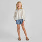 A young girl smiles with one foot forward, wearing a white long-sleeve blouse, Pink Chicken Girls' Scallop Short in chambray, and brown sandals against a plain background.