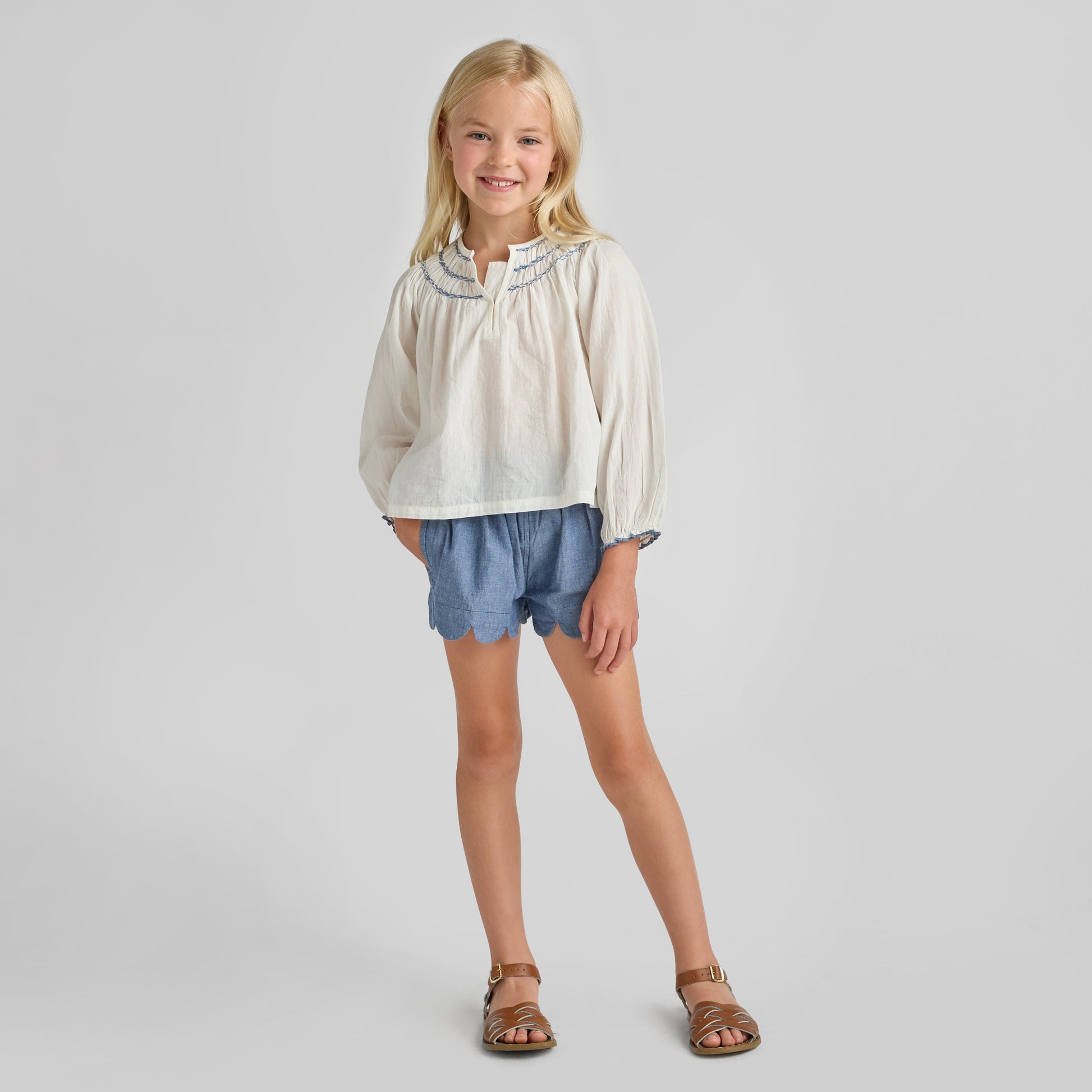 A young girl smiles with one foot forward, wearing a white long-sleeve blouse, Pink Chicken Girls' Scallop Short in chambray, and brown sandals against a plain background.