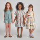 Three young girls stand in a row holding hands, smiling, and wearing Pink Chicken Girls’ Jules Shorts—patterned summer outfits featuring blue prairie blockprint, an elastic waist, and handy side pockets—set against a plain light gray background.