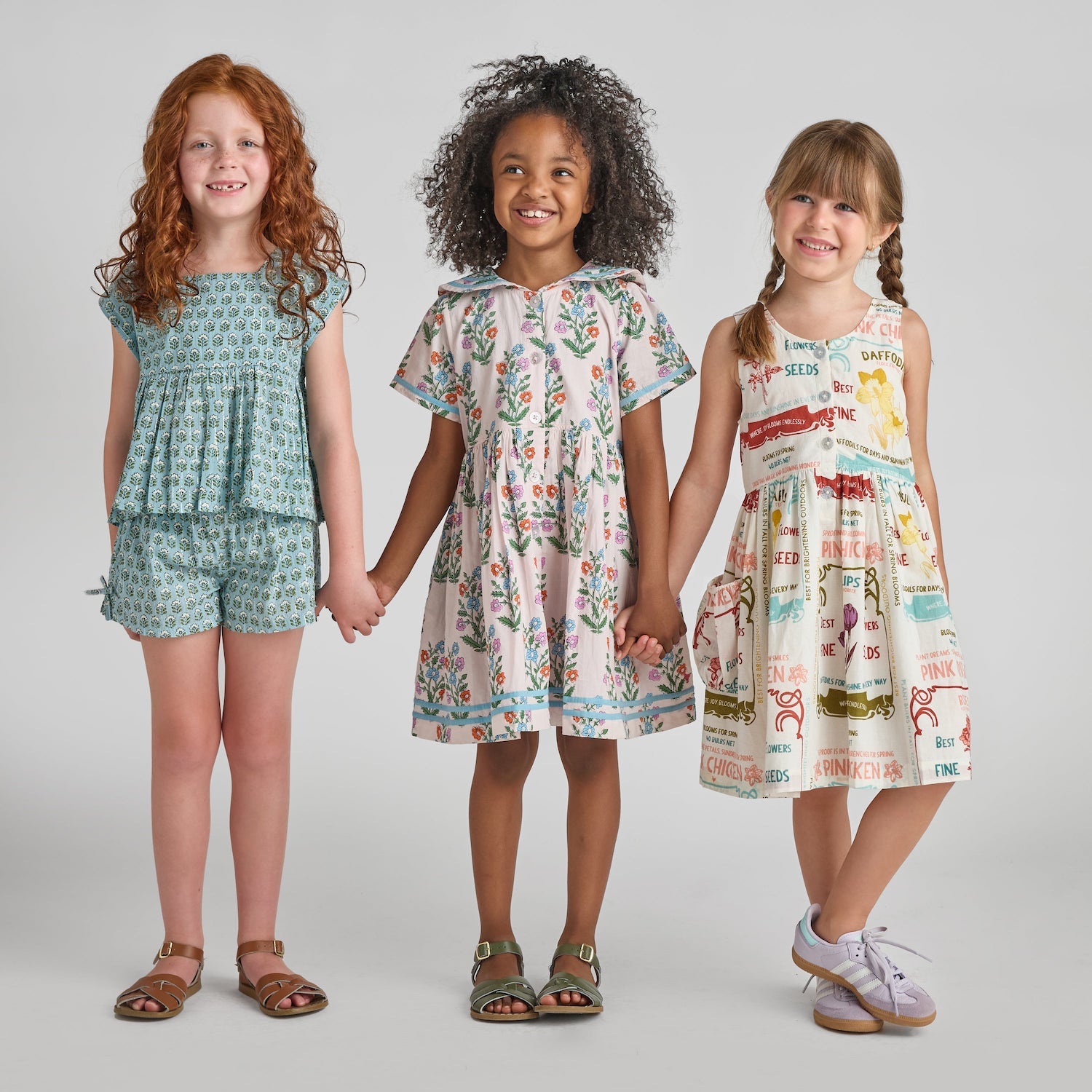 Three young girls stand in a row holding hands, smiling, and wearing Pink Chicken Girls’ Jules Shorts—patterned summer outfits featuring blue prairie blockprint, an elastic waist, and handy side pockets—set against a plain light gray background.