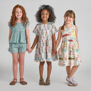 Three young girls stand in a row holding hands, smiling, and wearing Pink Chicken Girls’ Jules Shorts—patterned summer outfits featuring blue prairie blockprint, an elastic waist, and handy side pockets—set against a plain light gray background.
