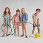 Four smiling children stand in a row with green garden hoses. One girl wears the Pink Chicken Girls' Autumn Dress, and a yellow watering can sits nearby on the floor against a plain gray background.