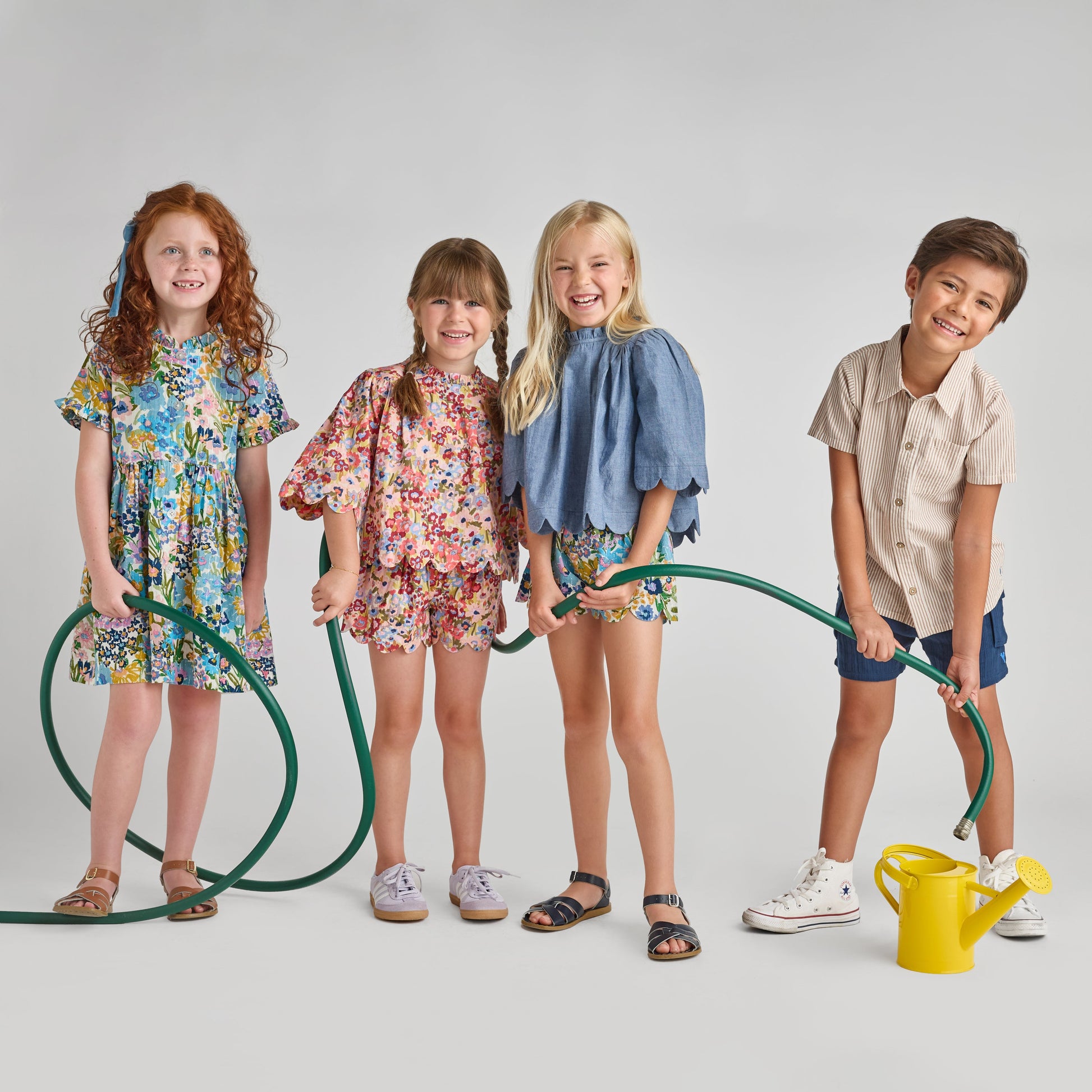 Four smiling children stand in a row with green garden hoses. One girl wears the Pink Chicken Girls' Autumn Dress, and a yellow watering can sits nearby on the floor against a plain gray background.