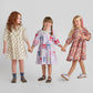Three young girls hold hands in different Pink Chicken Girls' Maribelle Dresses: one with dogs, one with colorful bows, and one in Burgundy Sketched Floral, all set against a plain background.