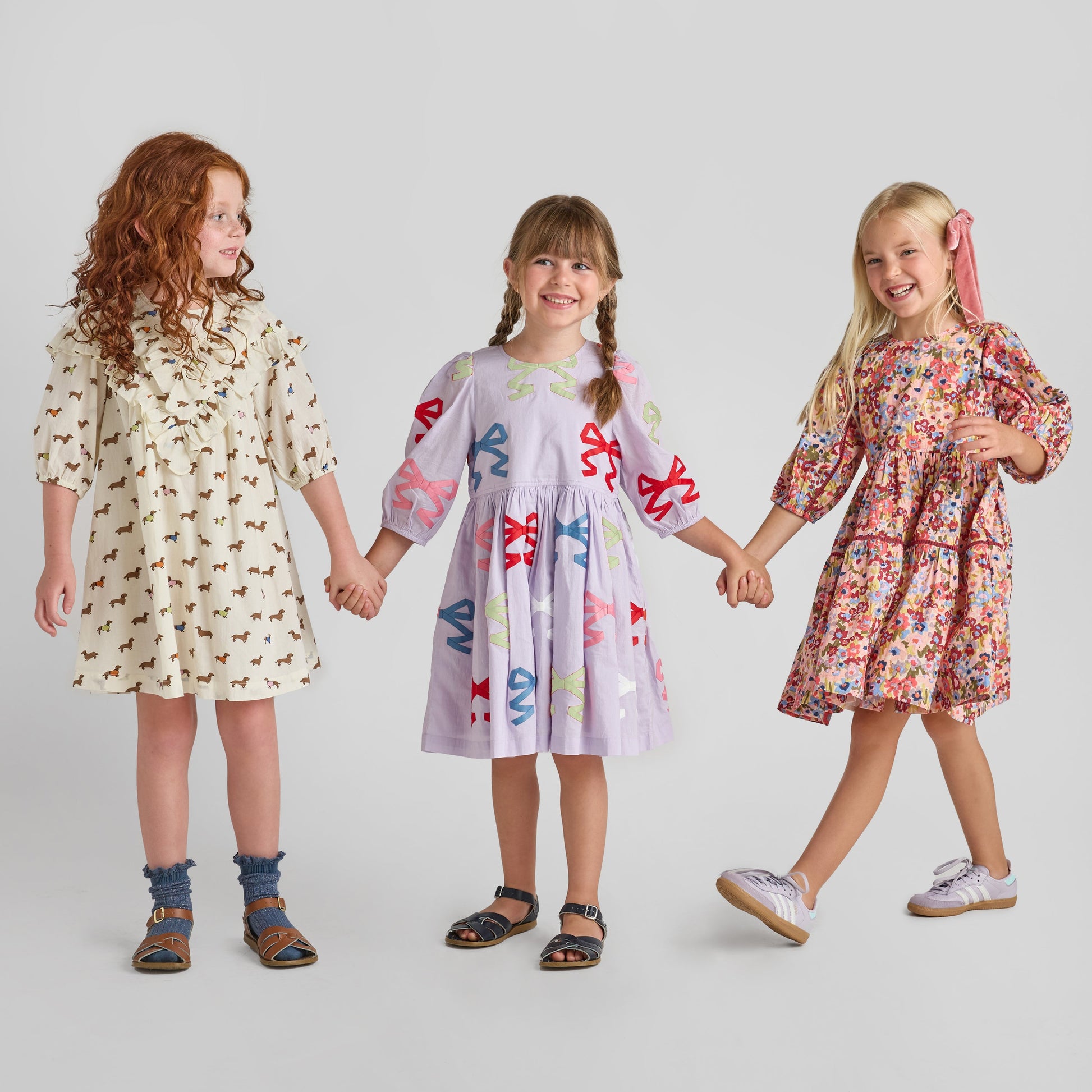 Three young girls hold hands in different Pink Chicken Girls' Maribelle Dresses: one with dogs, one with colorful bows, and one in Burgundy Sketched Floral, all set against a plain background.