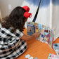A young girl with a red bow and checkered dress engages in imaginative play, arranging her Eeboo Paper Doll Set on the floor near a bookshelf and a small tent.