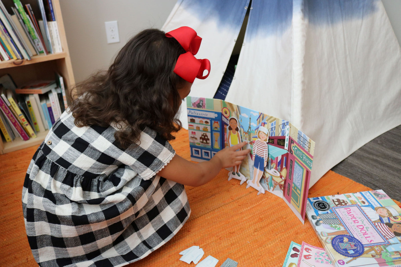 A young girl with a red bow and checkered dress engages in imaginative play, arranging her Eeboo Paper Doll Set on the floor near a bookshelf and a small tent.