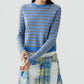 A person wearing the Pebble Stripe Shrunken Crew cashmere sweater by Guest in Residence—featuring a blue and brown stripe pattern—with a blue and yellow plaid shirt tied around their waist, stands against a plain light background.