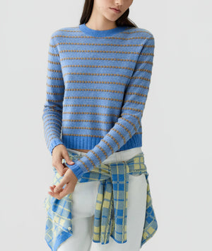 A person wearing the Pebble Stripe Shrunken Crew cashmere sweater by Guest in Residence—featuring a blue and brown stripe pattern—with a blue and yellow plaid shirt tied around their waist, stands against a plain light background.