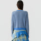 A person with long dark hair, seen from behind, wearing the Pebble Stripe Shrunken Crew by Guest in Residence—a blue and brown striped cashmere sweater—paired with a layered blue and yellow plaid skirt, styled with a cropped fit against a plain light background.