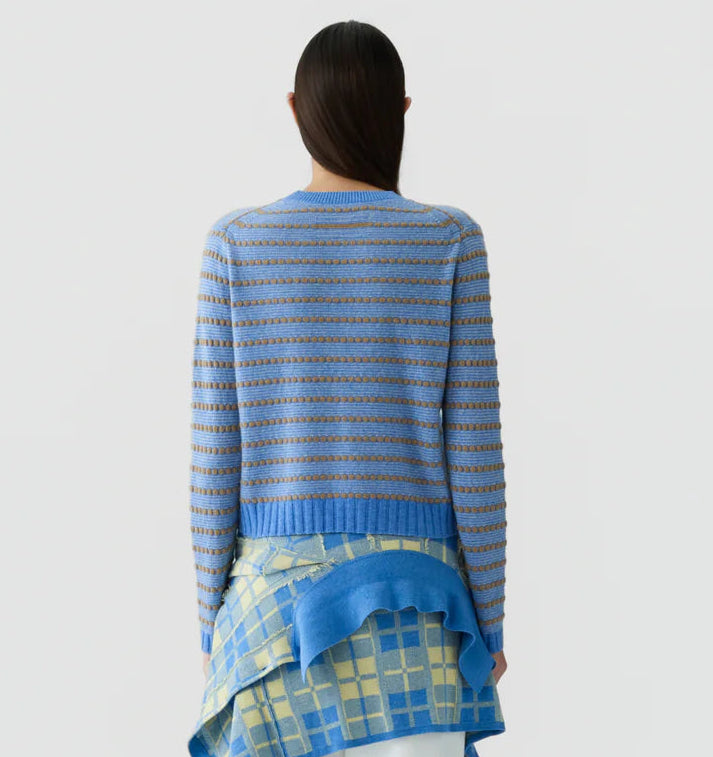 A person with long dark hair, seen from behind, wearing the Pebble Stripe Shrunken Crew by Guest in Residence—a blue and brown striped cashmere sweater—paired with a layered blue and yellow plaid skirt, styled with a cropped fit against a plain light background.