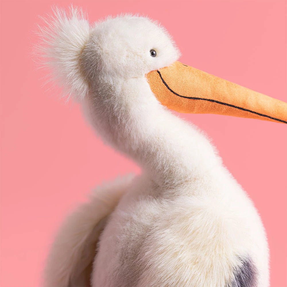 The Jellycat Keli Pelican, a white plush toy with an orange beak inspired by "The Beak" talk show, is displayed against a solid pink background.