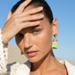 A person with light skin and dark hair poses with a hand on their forehead, wearing Lizzie Fortunato Raffia Earrings and a crocheted top against a blue sky background. Their jet-setter personality shines through.