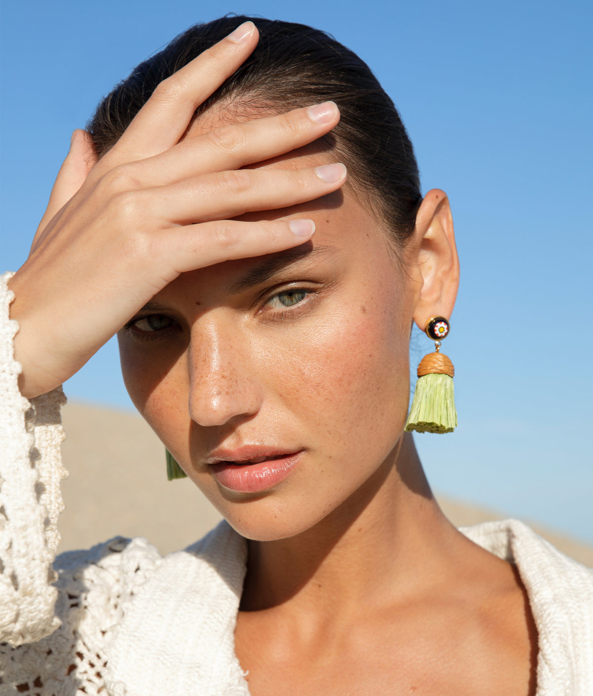 A person with light skin and dark hair poses with a hand on their forehead, wearing Lizzie Fortunato Raffia Earrings and a crocheted top against a blue sky background. Their jet-setter personality shines through.