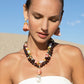 A woman adorned with Lizzie Fortunato Raffia Earrings and multi-colored beaded necklaces stands against a clear sky backdrop. She gazes downward, donning a white top that exudes a jet-setter personality.