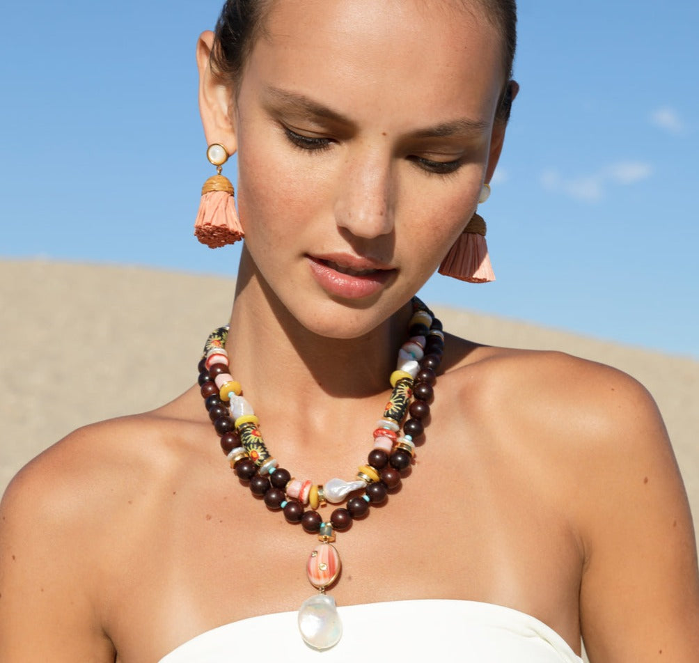 A woman adorned with Lizzie Fortunato Raffia Earrings and multi-colored beaded necklaces stands against a clear sky backdrop. She gazes downward, donning a white top that exudes a jet-setter personality.