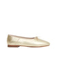 The Dream Ballerina by Mansur Gavriel is a gold metallic flat in Italian lambskin leather with a knotted bow, beige trim, and a flat brown sole, shown in side profile against a white background.