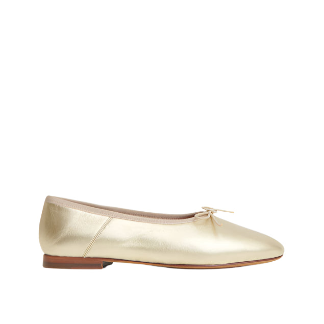 The Dream Ballerina by Mansur Gavriel is a gold metallic flat in Italian lambskin leather with a knotted bow, beige trim, and a flat brown sole, shown in side profile against a white background.