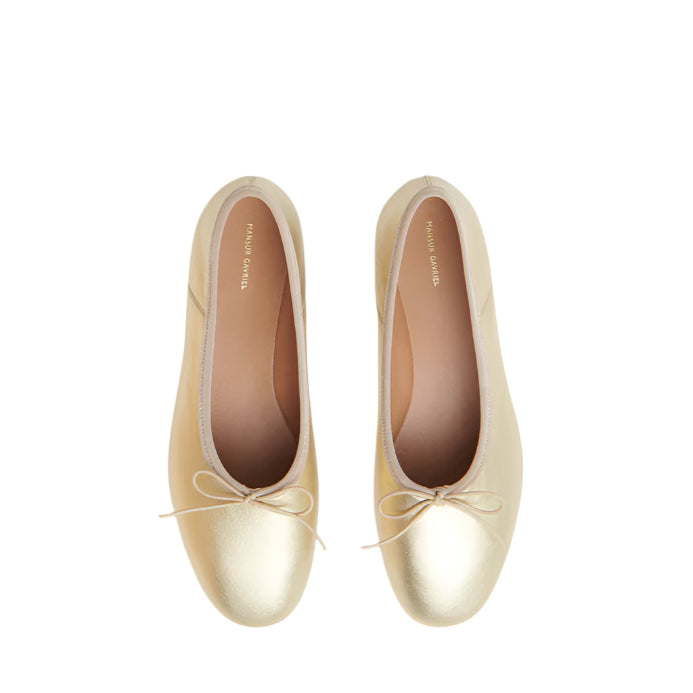 The Dream Ballerina by Mansur Gavriel is a pair of gold Italian lambskin flats featuring a delicate knotted bow on the toes, shown from above against a white background.
