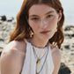A woman with long red hair and freckles wears a sleeveless white top, layered necklaces, and Lizzie Fortunato Minnow Hoop Earrings, sitting outdoors near rocks.