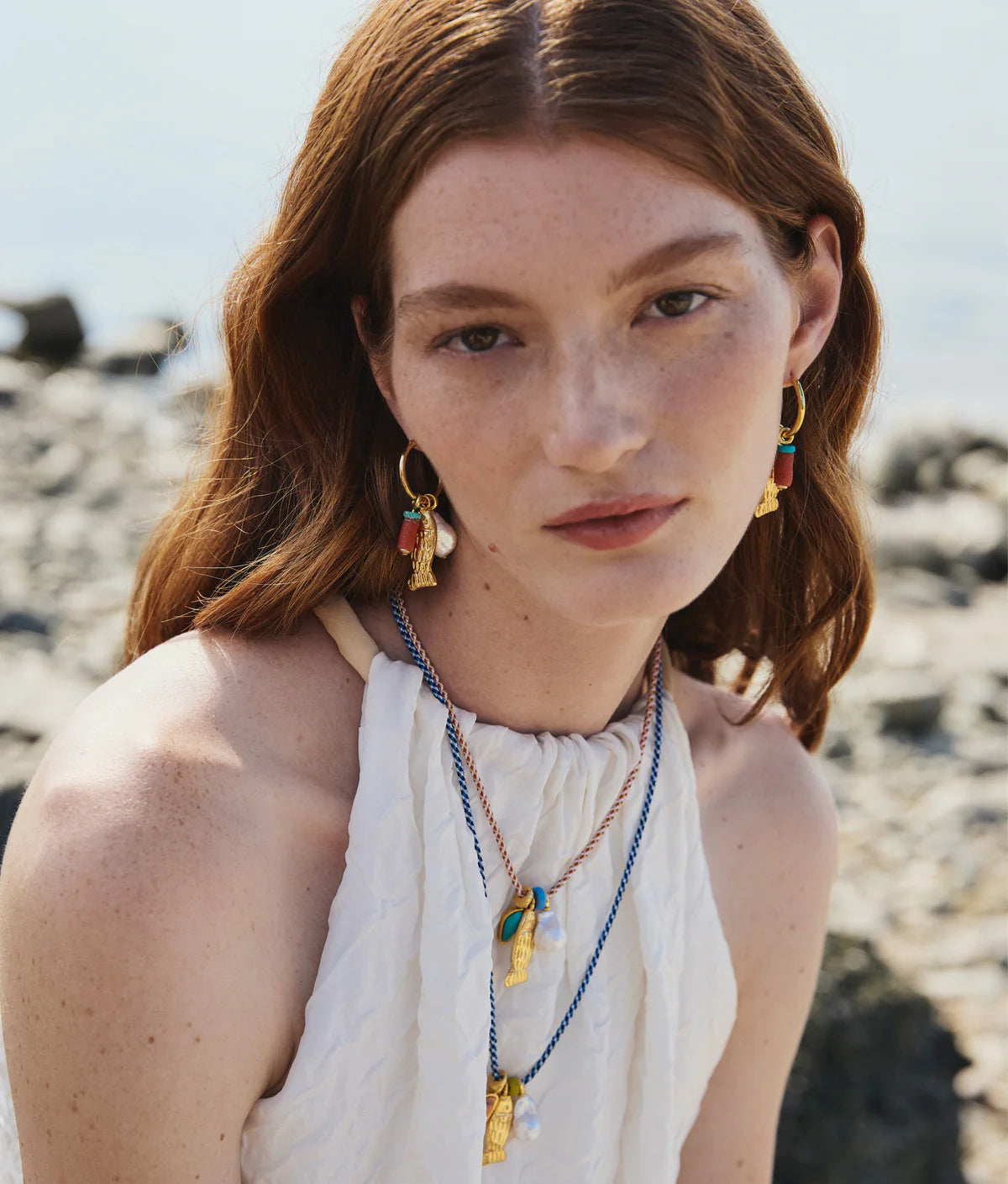 A woman with long red hair and freckles wears a sleeveless white top, layered necklaces, and Lizzie Fortunato Minnow Hoop Earrings, sitting outdoors near rocks.