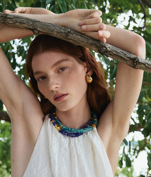 A person with long red hair stands outdoors, wearing a sleeveless white top and the Lizzie Fortunato Toga Beach Necklace, along with earrings, resting their arms on a tree branch.