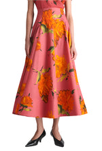 A person wearing the Lela Rose Parker Floral Skirt, a pink midi with bold orange blooms and a high waist, styled with black low-heeled shoes. The photo is cropped from the shoulders down.