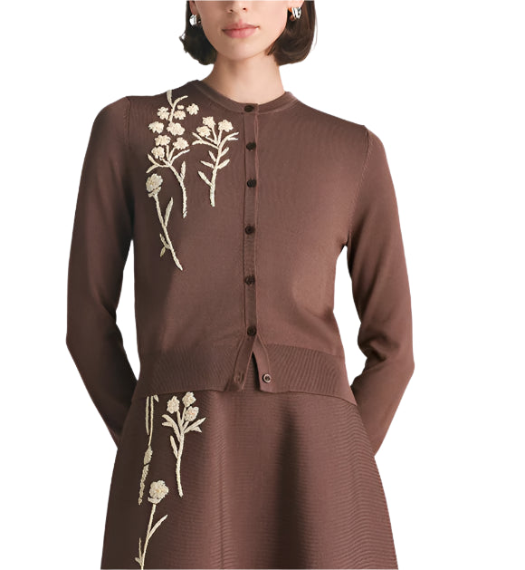A woman wears the Lela Rose Raffia Embroidered Knit Long Sleeve Cardigan, a brown piece with white floral embroidery on the left side, standing against a white background.
