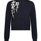 The Lela Rose Raffia Embroidered Knit Long Sleeve Cardigan features a navy blue knit fabric, button-up front, and white floral embroidery on the left shoulder and chest.