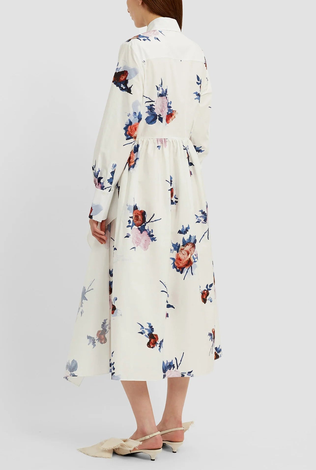 A woman is shown from behind wearing the Erdem Long Sleeve Midi Shirt Dress in white with a rose bouquet print, paired with beige pointed-toe, low-heeled shoes.