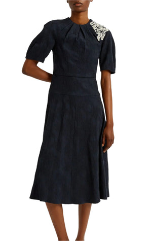 A person wearing the Erdem Short Sleeve Midi Dress, a navy blue, knee-length fit-and-flare style featuring short sleeves and a white floral applique on the left shoulder.