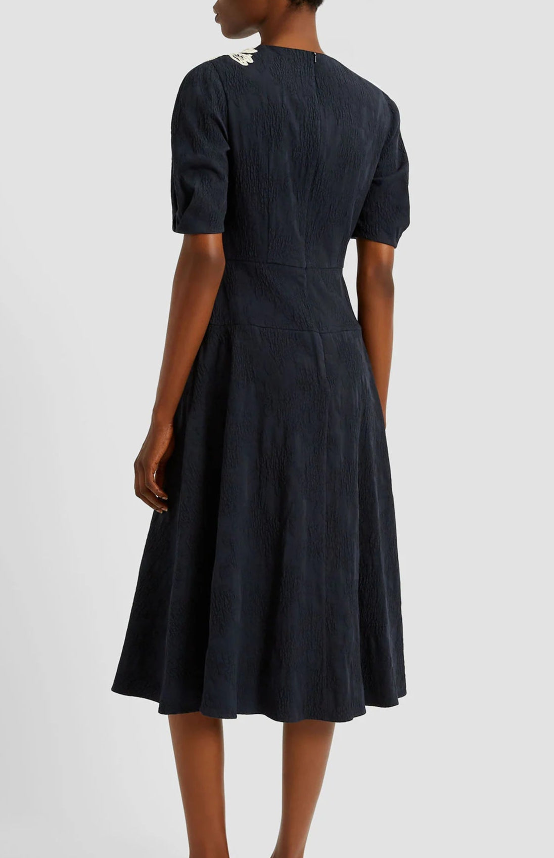A person stands facing away, wearing the Erdem Short Sleeve Midi Dress in navy jacquard, featuring a knee-length silhouette and a small white detail on the shoulder.