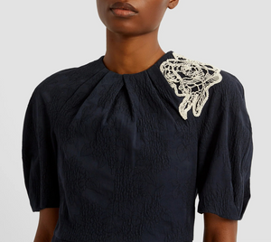 A person wearing the Erdem Short Sleeve Midi Dress—a navy jacquard style with white lace floral applique on one shoulder.