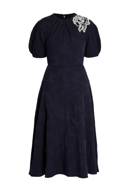The Erdem Short Sleeve Midi Dress features a knee-length navy jacquard design with short puff sleeves, subtle textured pattern, and a white floral applique accenting one shoulder.
