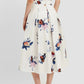 A person stands facing away, wearing Erdem's Painted Rose Bouquet Pleated Midi Skirt with a blue and red floral design, a matching top, and beige pointed-toe flats with bows.