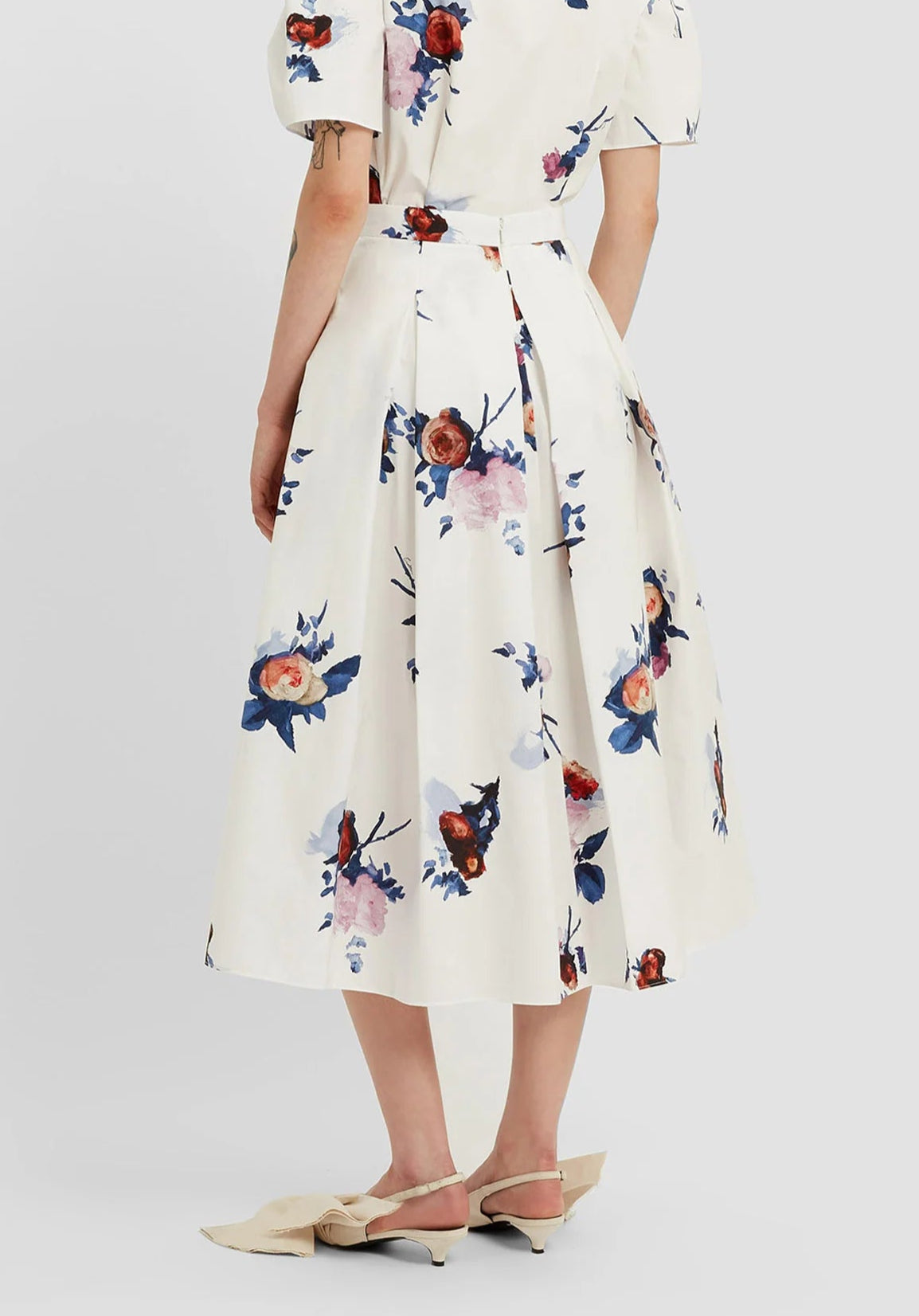 A person stands facing away, wearing Erdem's Painted Rose Bouquet Pleated Midi Skirt with a blue and red floral design, a matching top, and beige pointed-toe flats with bows.
